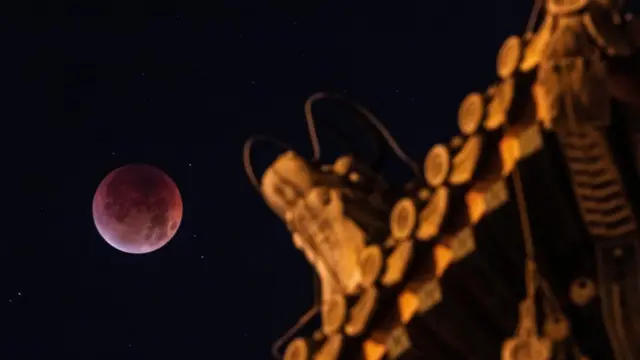 Një hënë me ngjyrë të kuqe në qiellin e natës, përpara saj është një pjesë e çatisë së portës Yongdingmen me një statujë qilin.